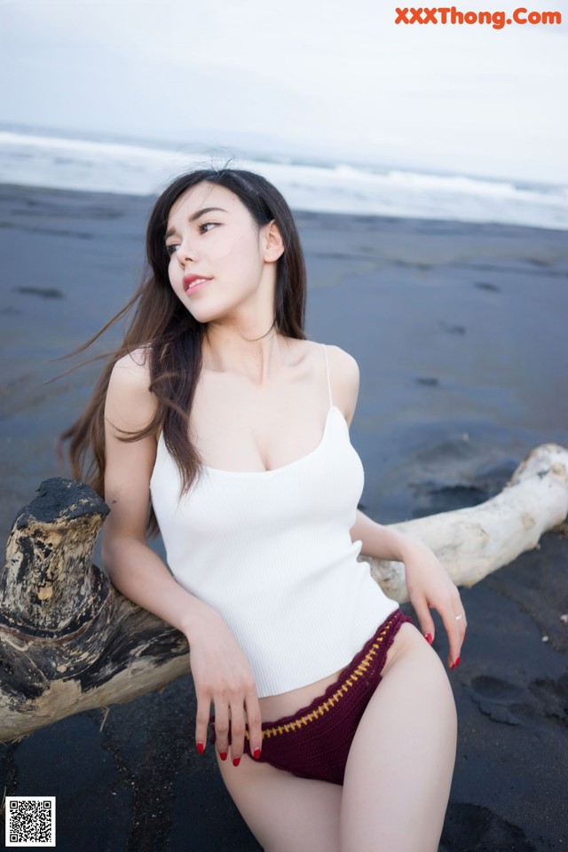 A woman in a white tank top sitting on a log on the beach.