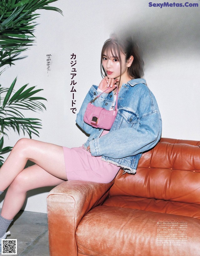 A woman sitting on a brown couch wearing a denim jacket and pink skirt.