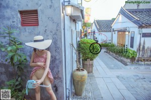 A woman in a pink dress and a straw hat is standing on a sidewalk.