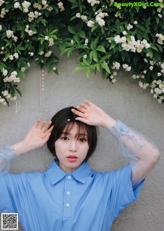 A woman in a blue shirt is posing in front of a wall of flowers.