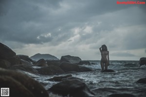 A naked woman standing on a rock by the ocean.